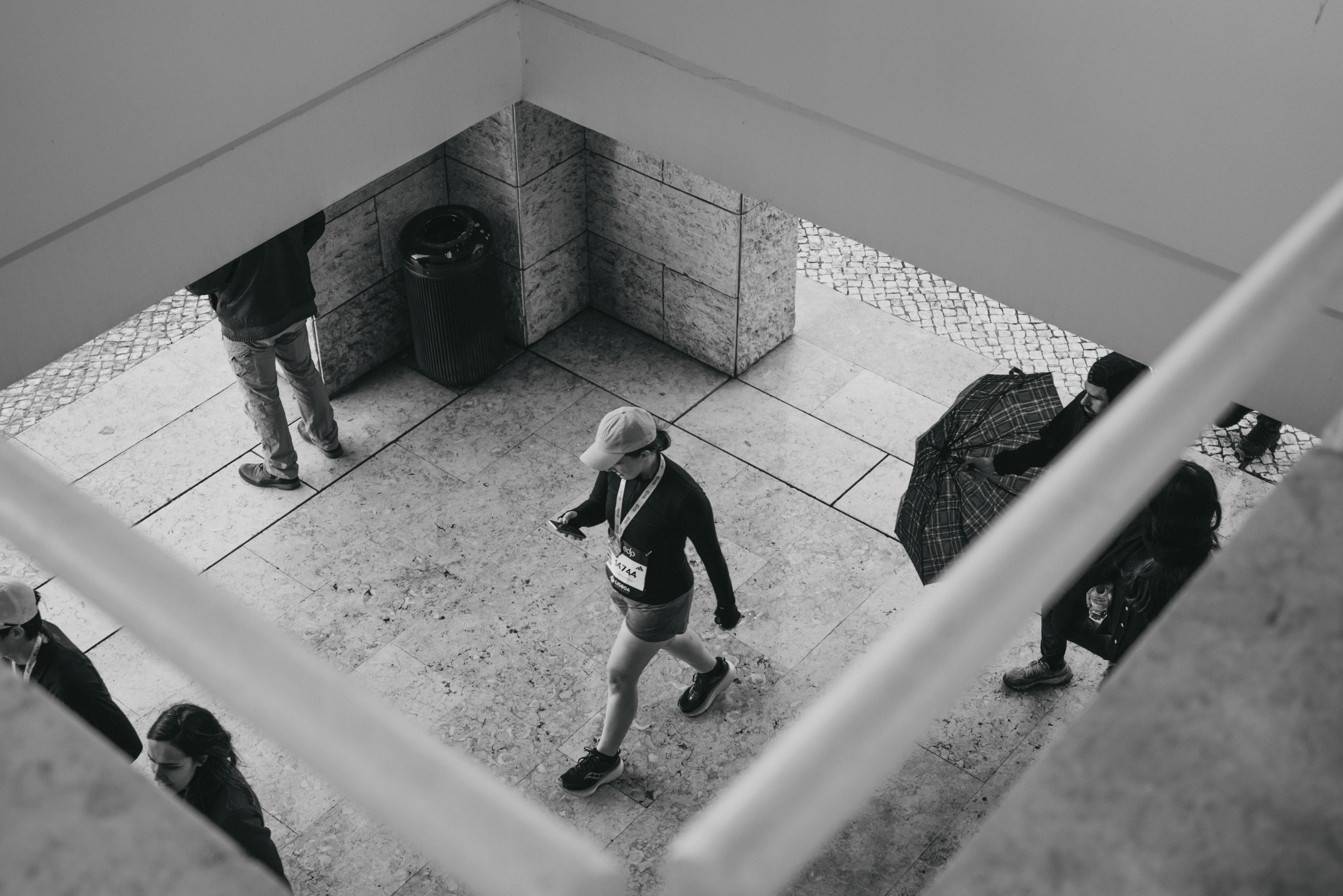 Black and white overhead view through a stairwell railing, showing a woman wearing a race bib and medal walking across a marble courtyard, with other people nearby.
