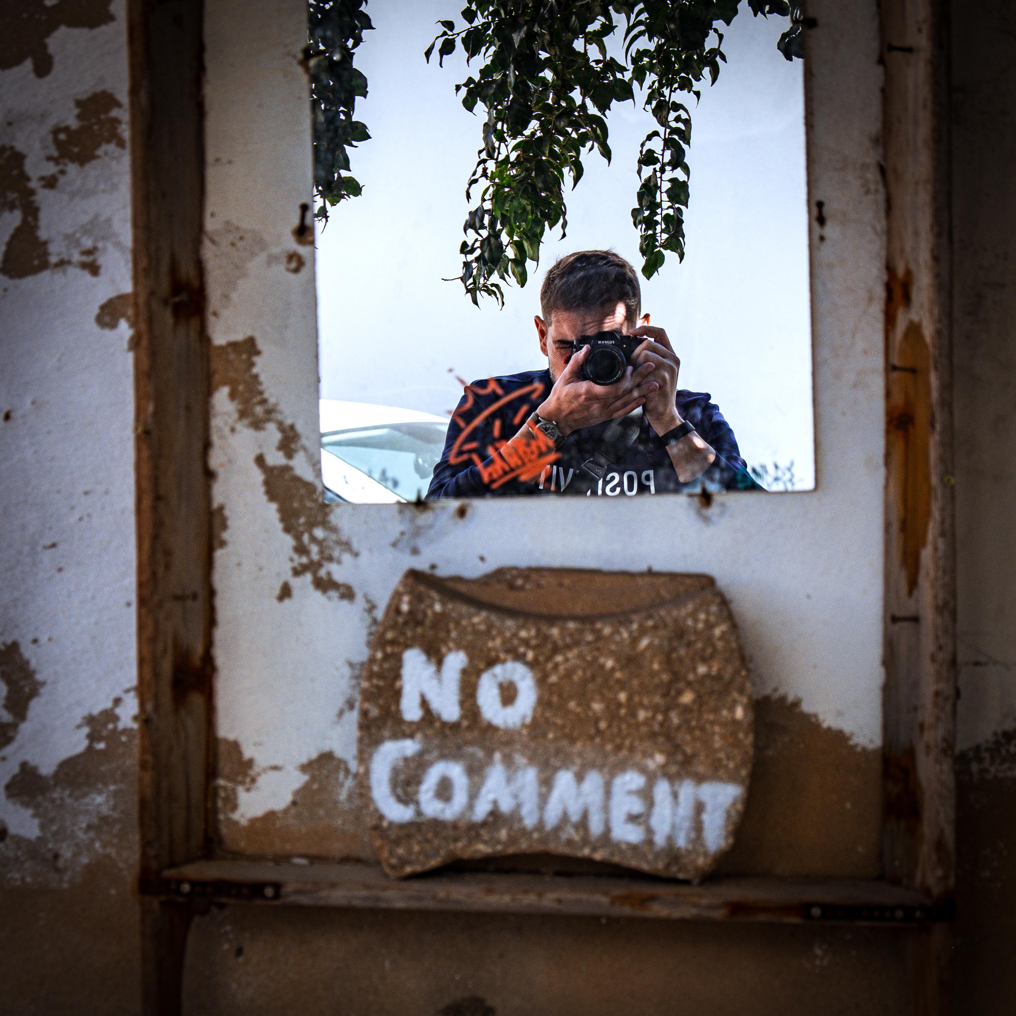 Self-portrait reflected in a weathered mirror mounted on a peeling wall, showing a man photographing himself with a Fujifilm camera, hanging ivy above and a stone tile reading 'NO COMMENT' below.