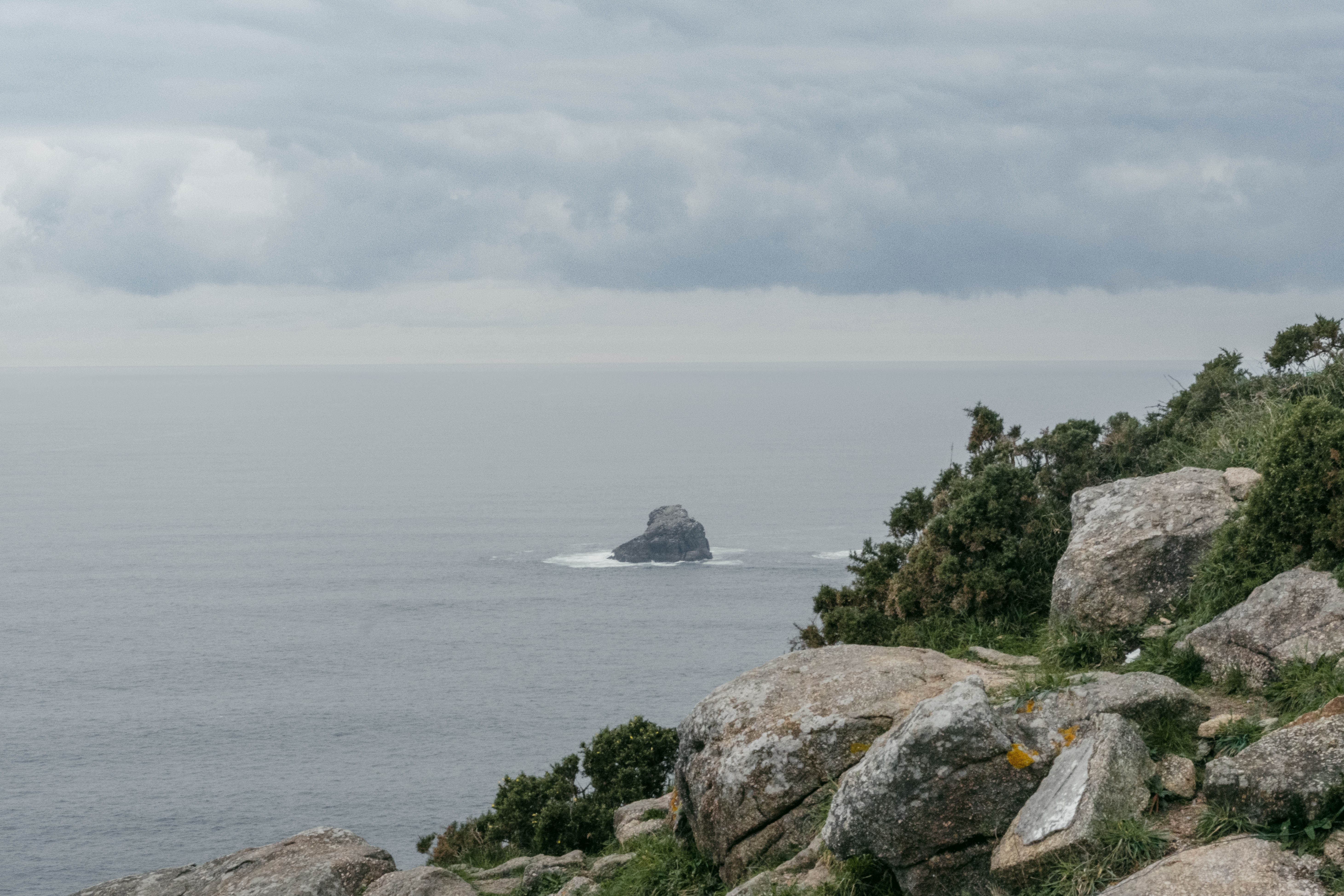 View from a rocky, gorse-covered cliff overlooking a calm grey sea, with a solitary rock formation surrounded by waves in the distance, under an overcast sky