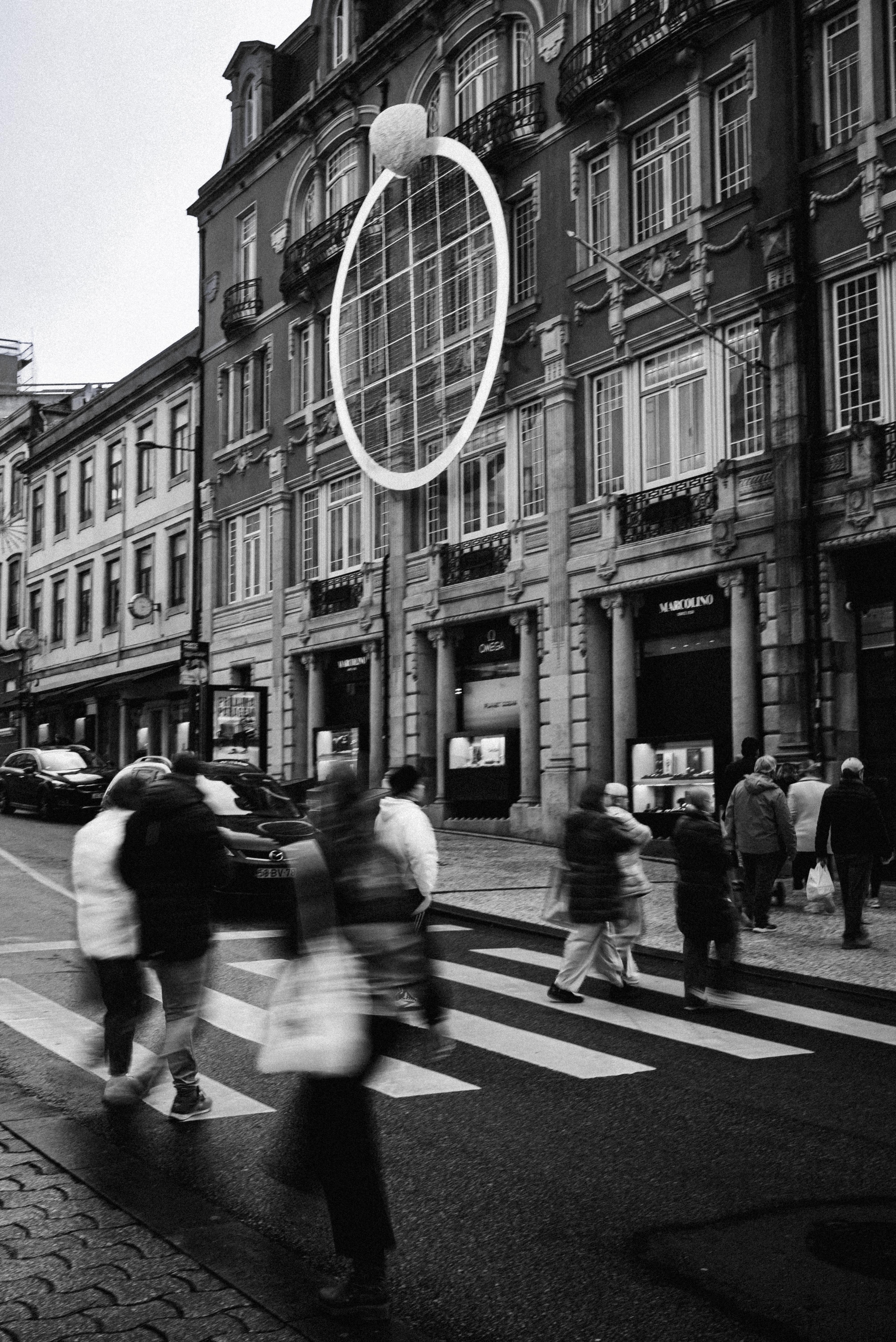 Black and white street photograph of pedestrians crossing a city street with motion blur, beneath a large ring-shaped sculpture, with ornate historic building facades and jewelry shop windows in the background.