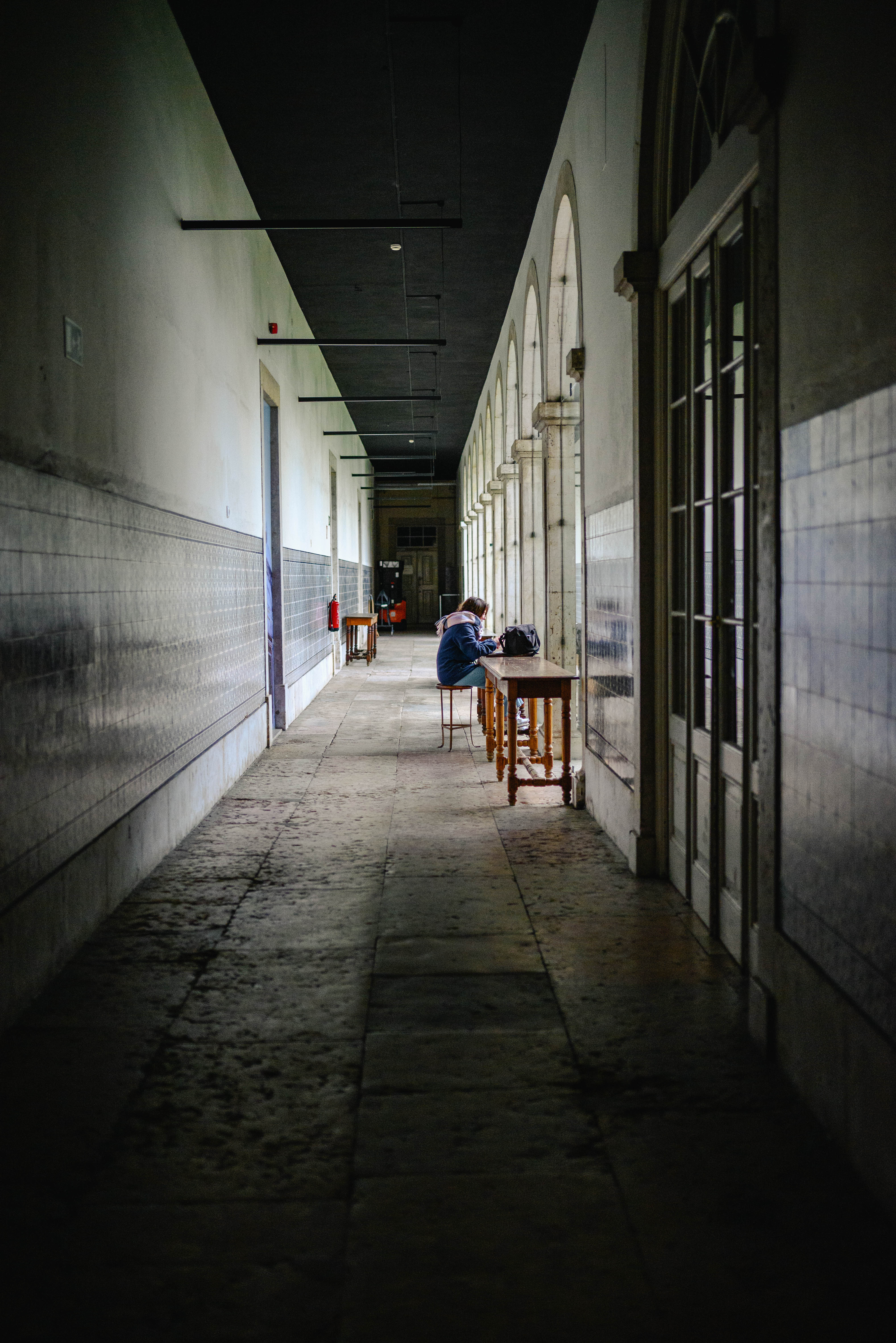 A long, symmetrical perspective shot of a quiet, lengthy arcade flanked by solid walls, flooded with natural light from tall windows, the scene culminates in a distant subject seated at a table, bathed in natural light.