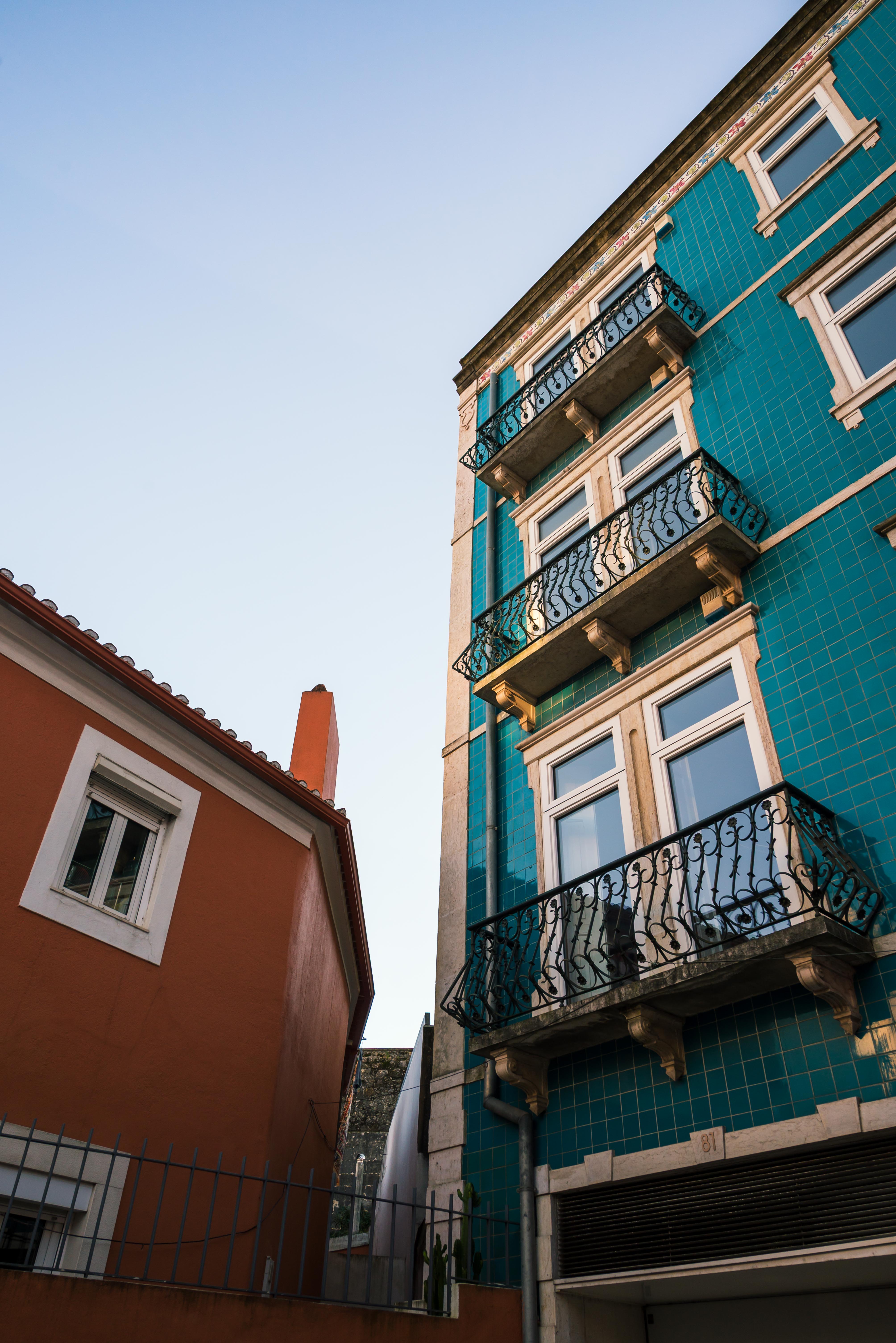 Low-angle view of a teal azulejo-tiled building with ornate wrought-iron balconies, standing beside a terracota-colored house, against a clear blue sky.