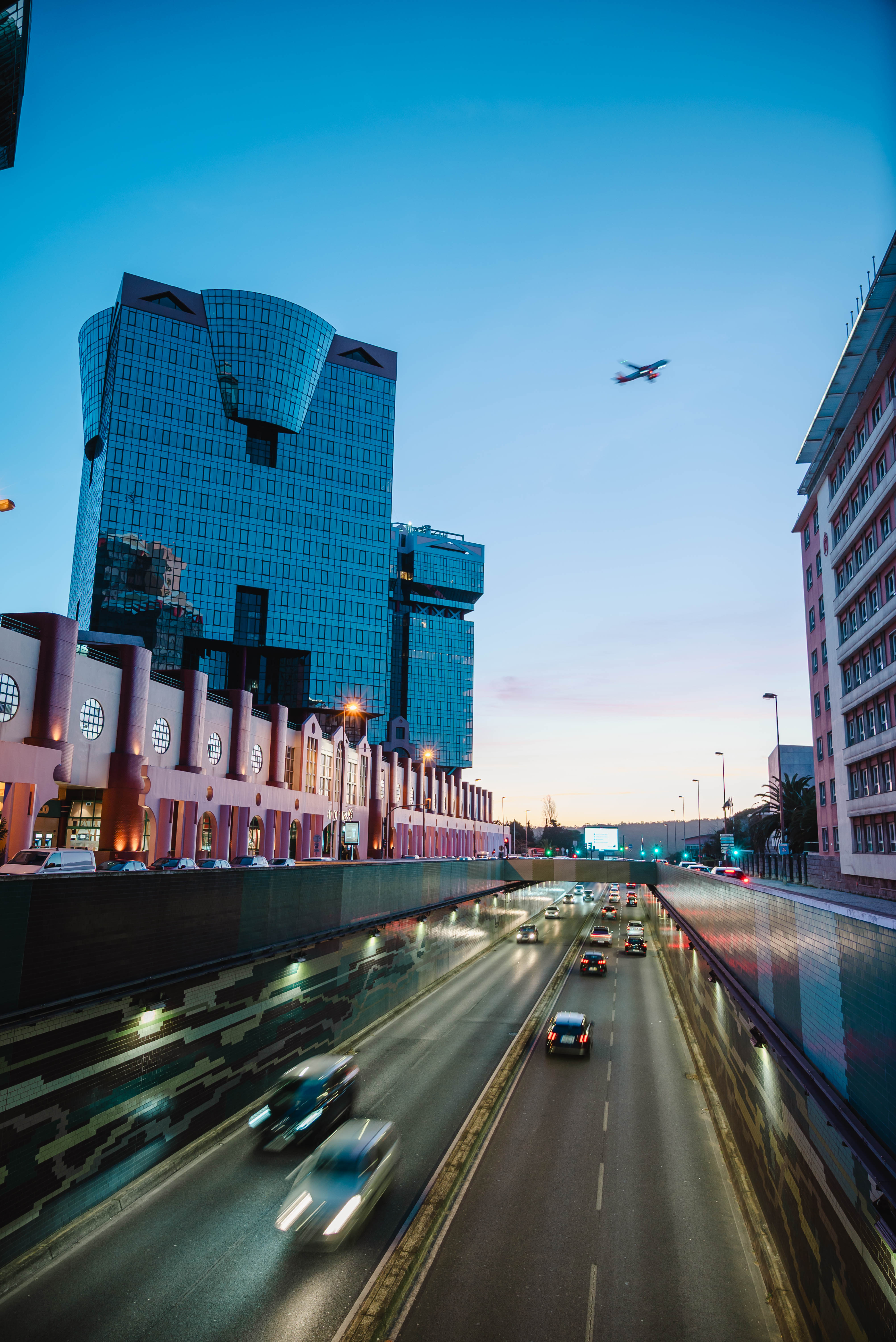 Blue hour highway tunnel in Lisbon, with the postmodern Amoreiras building (blue glass towers, salmon-pink façade) in the background and a plane in the sky.