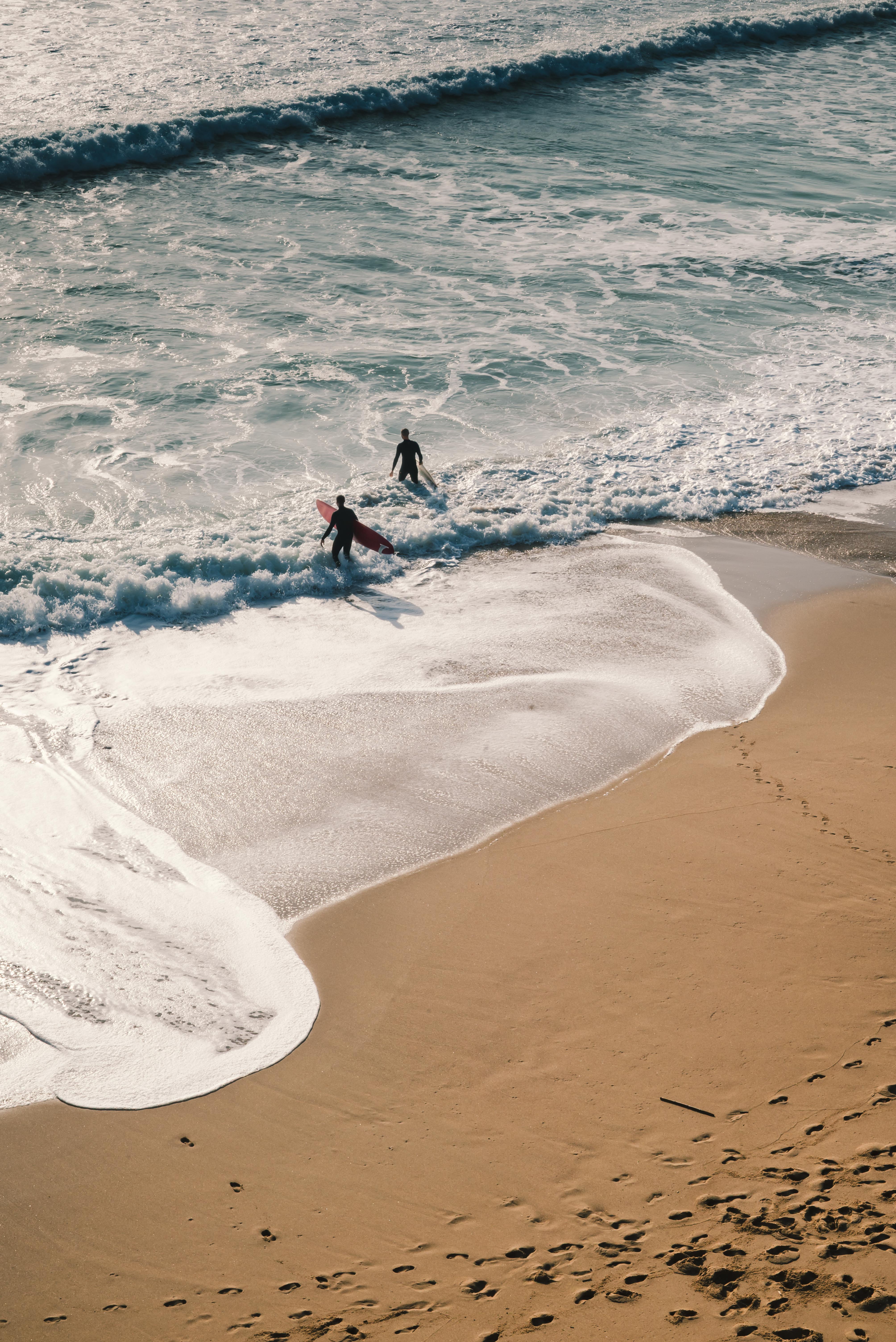 High aerial perspective view of two surfers walking with their boards onto the cresting waves at a wide sandy beach. The foam patterns spread across the sand like natural maps.