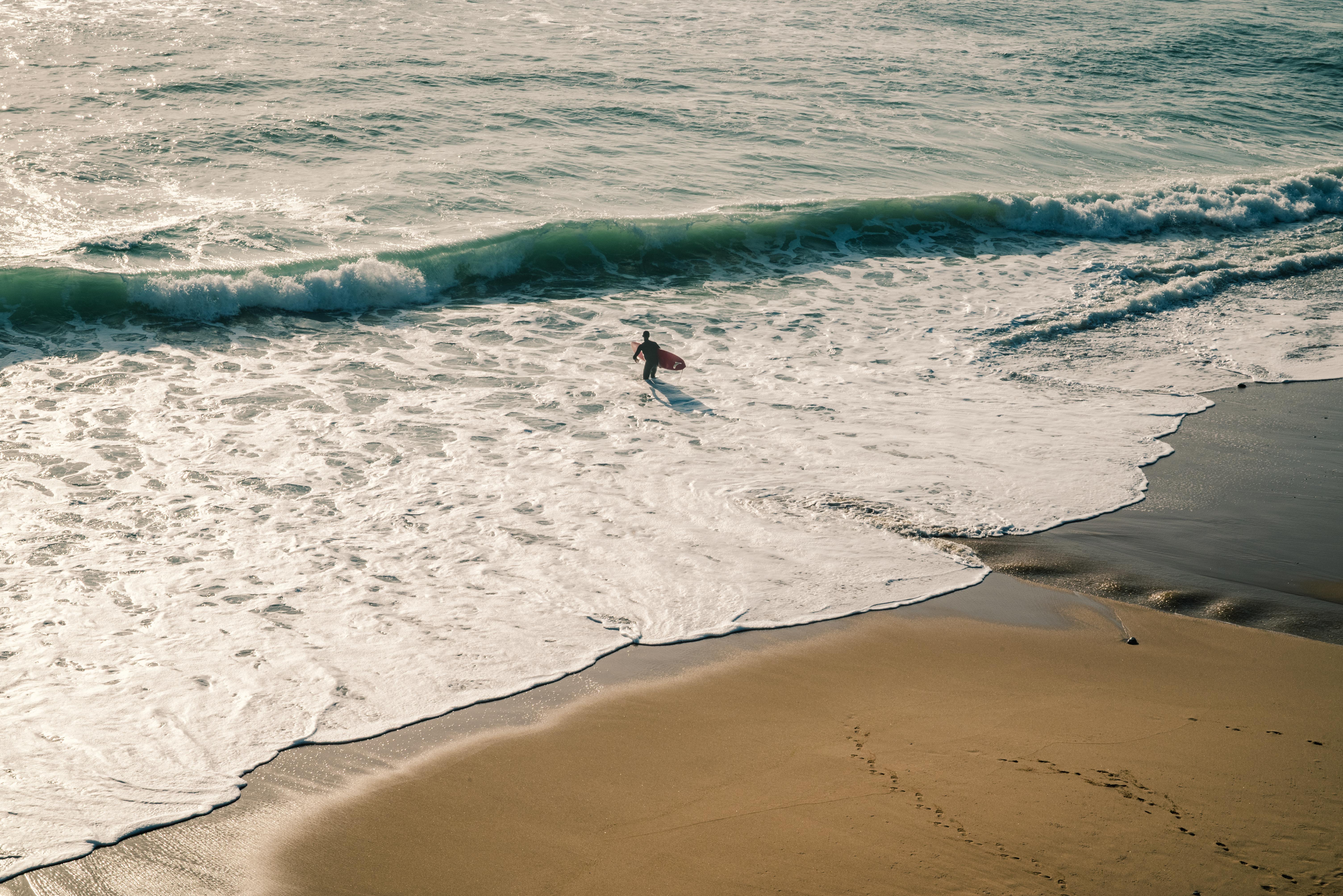High-angle view of the ocean, showing foam cresting around a single surfer as they ride a wave toward the shore, flanked by wet sand and foam.