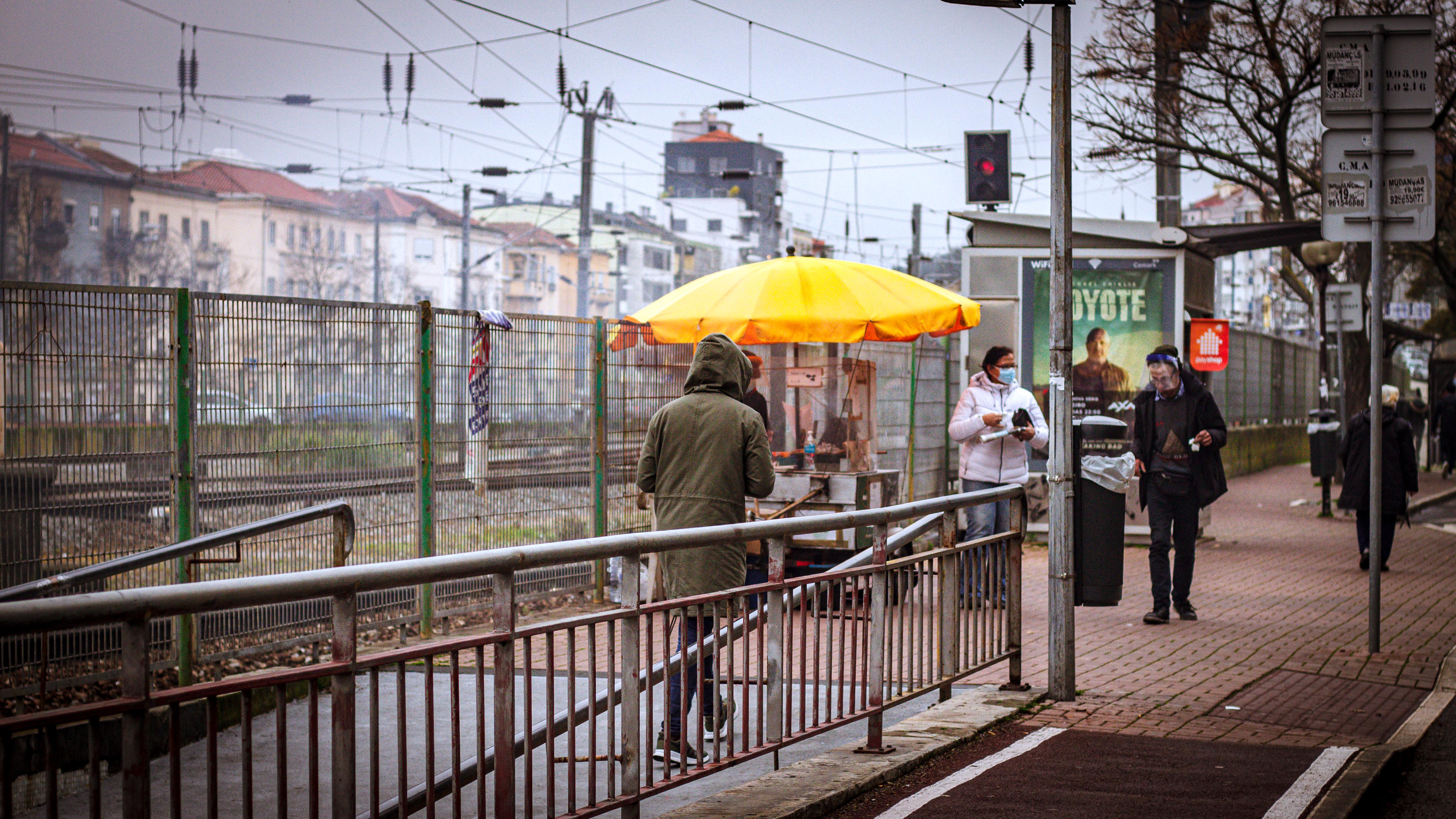 Street photograph of a food cart with a bright yellow umbrella beside a railway fence, with pedestrians wearing face masks standing nearby on a grey overcast day.
