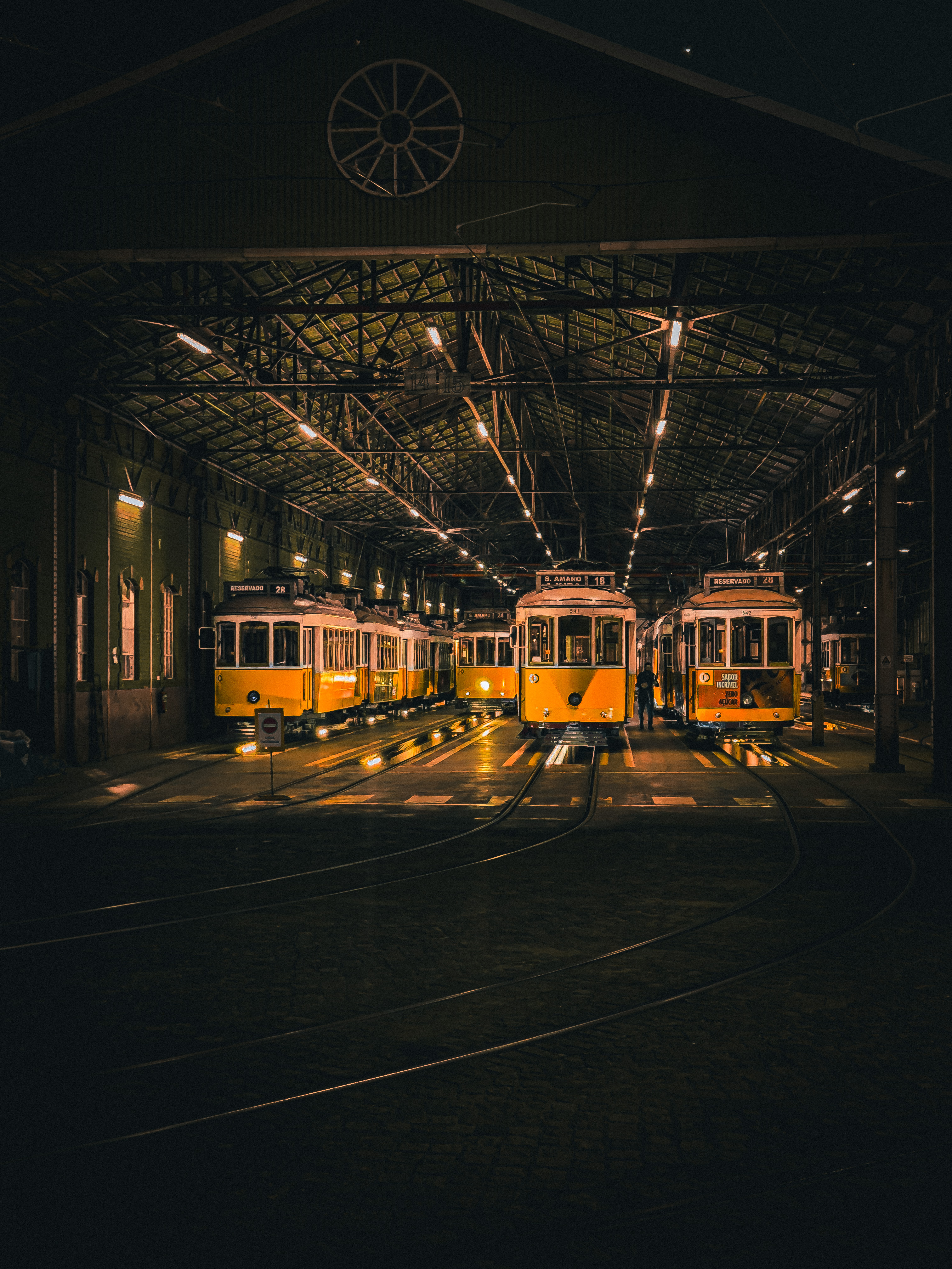 Night interior of a tram depot with several yellow Lisbon trams parked in rows under warm overhead lighting, tracks curving across the foreground floor."