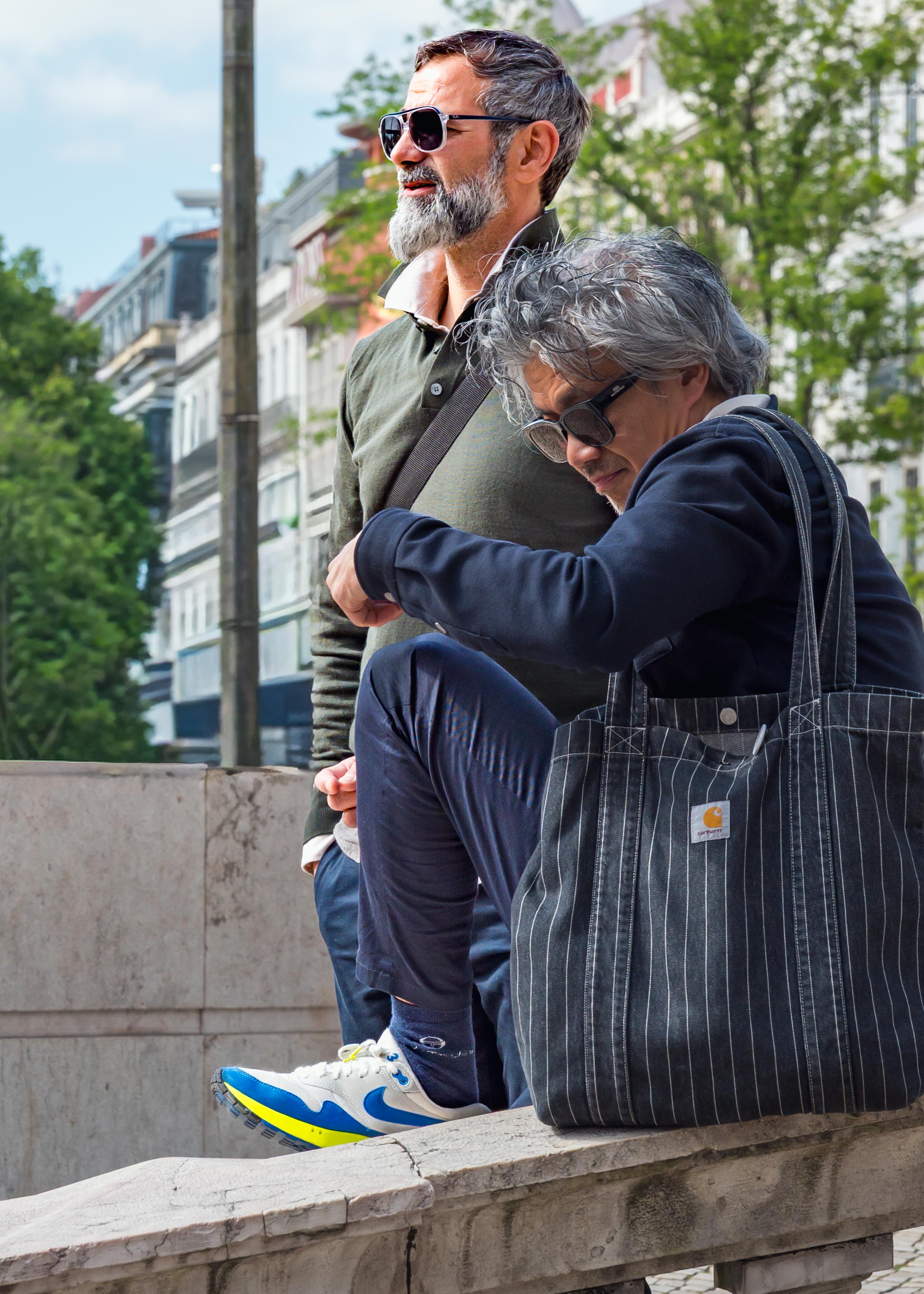 Street photograph of two men sitting on a stone wall in a city, one wearing sunglasses and a green polo shirt looking off into the distance, the other checking his watch, with a striped tote bag beside him and leafy trees and buildings in the background.
